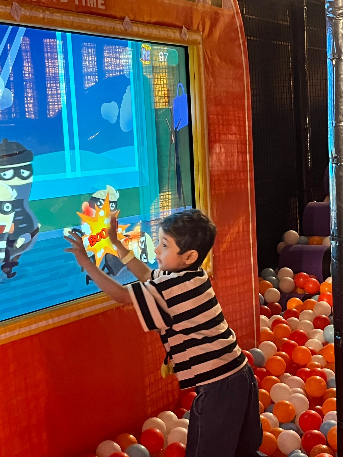 A young boy plays an interactive game, reaching out to a screen with cartoon characters, surrounded by a ball pit.