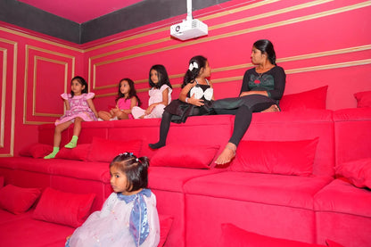 A group of young girls and a woman sit on a bright red couch in a pink room, watching something.