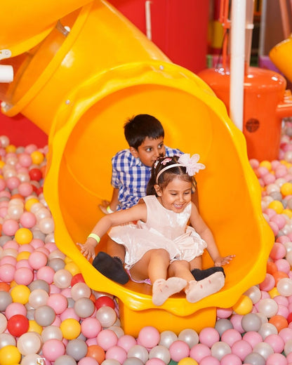 A young girl slides down a yellow tube into a ball pit, with a boy behind her.