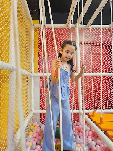 A young girl in a blue outfit plays in a ball pit, holding onto ropes in an indoor playground.