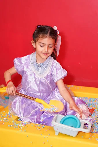 A young girl in a purple dress plays with colorful beads in a yellow sandbox.
