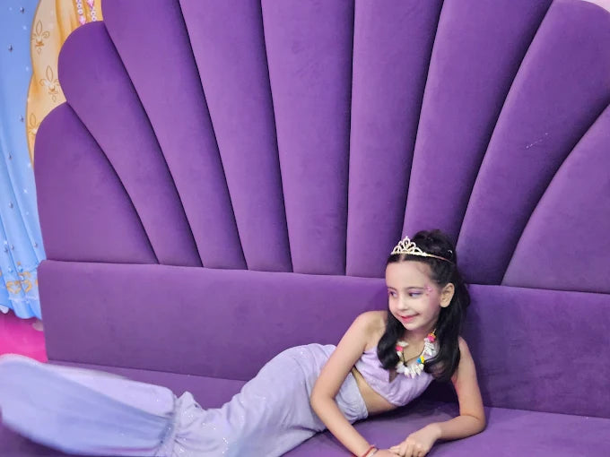 A young girl in a mermaid costume lies on a purple seashell-shaped couch. She wears a tiara and a shell necklace.