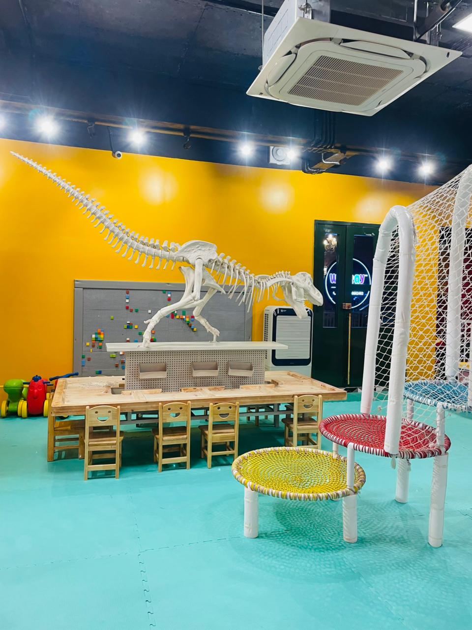 A dinosaur skeleton is displayed in a brightly colored children's play area with small tables and chairs.
