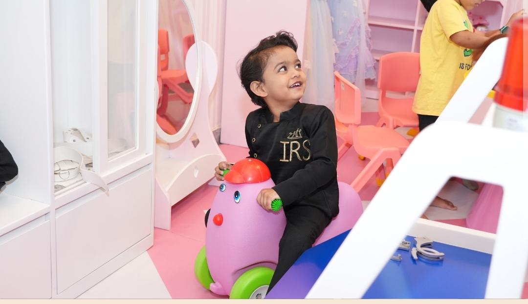 A young boy in a black outfit rides a pink toy car in a brightly colored room.