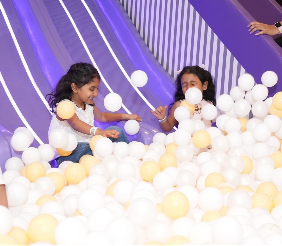 Two young girls play in a ball pit with a purple slide.