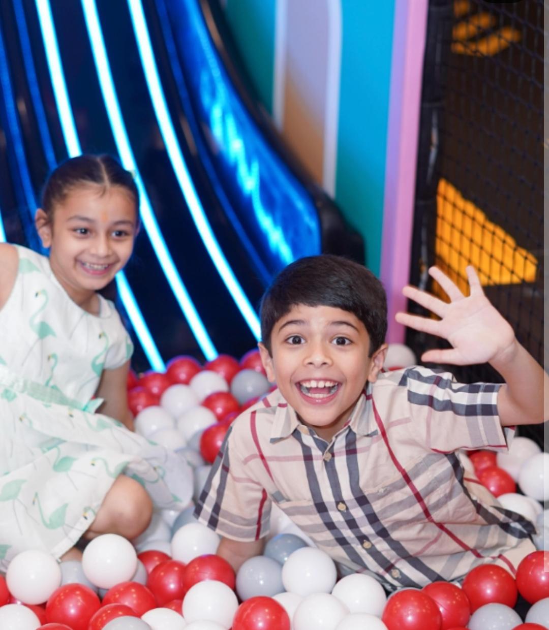 Two smiling children play in a ball pit with red, white, and gray balls.