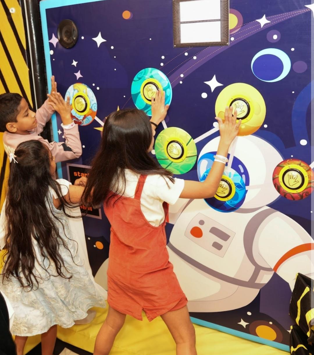 Children interact with a space-themed game wall featuring colorful buttons and an astronaut graphic.