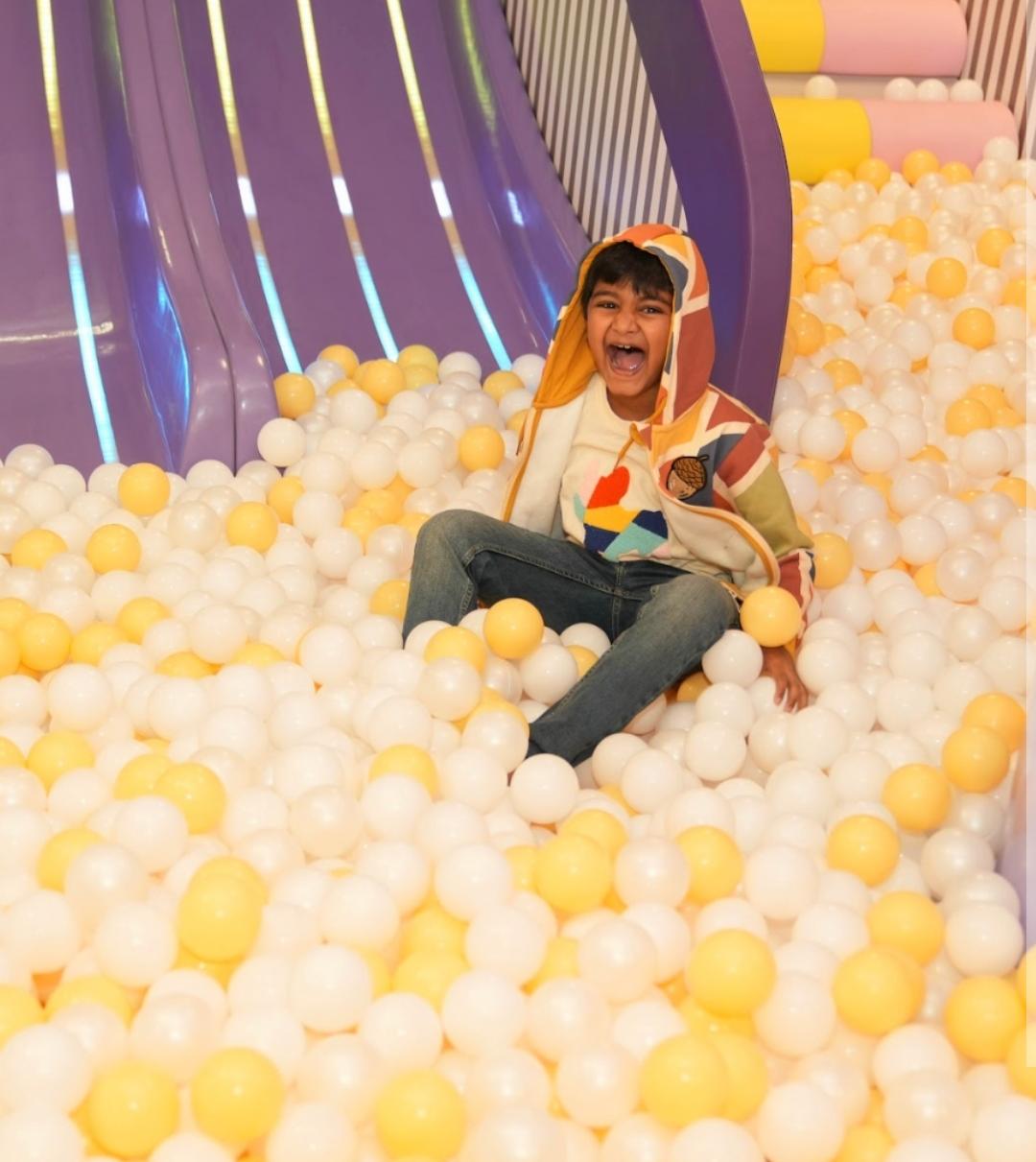 A young child laughs joyfully while sitting in a ball pit with a purple slide in the background.