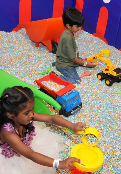 Two young children play with toy trucks and balls in a ball pit.