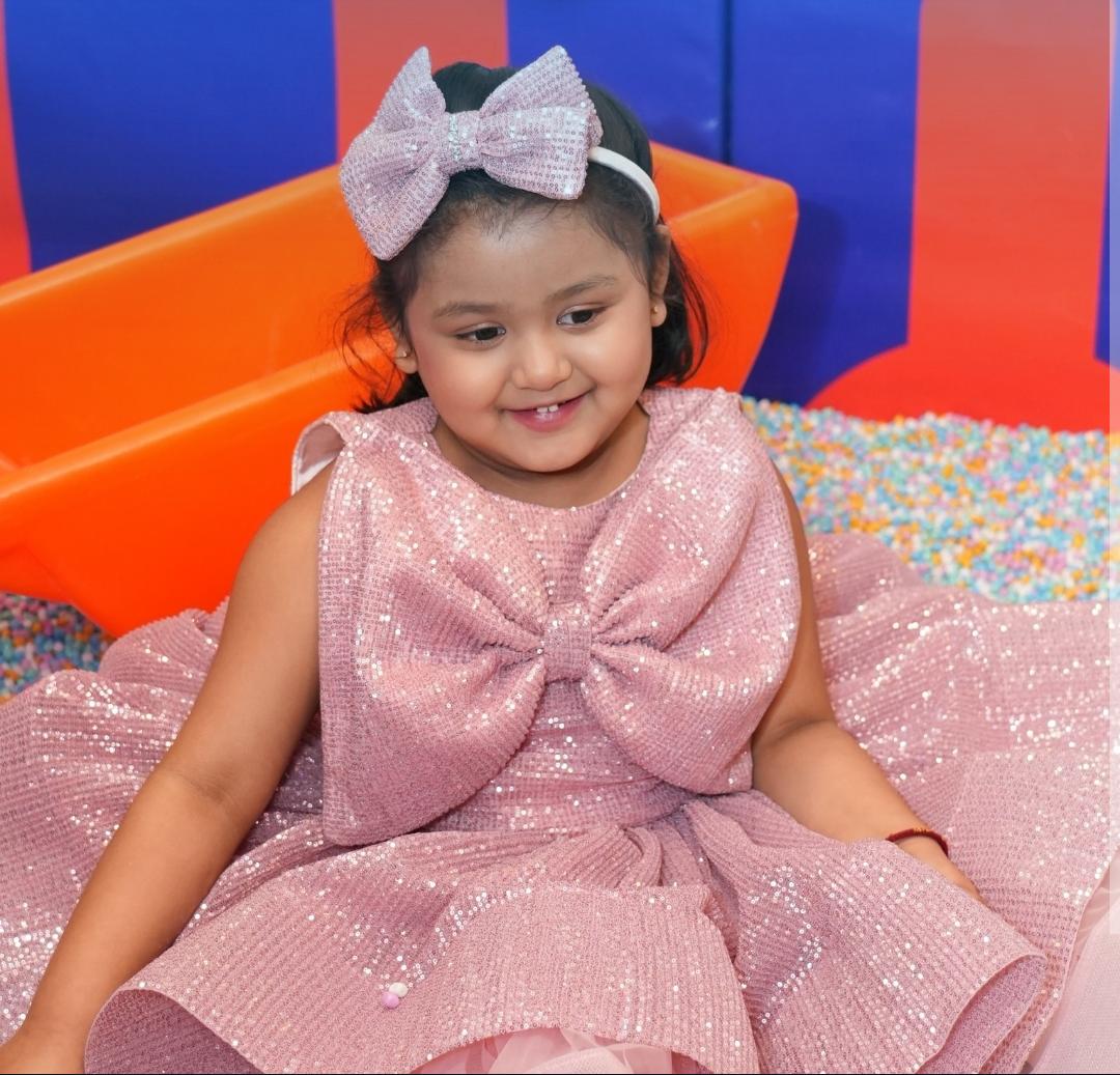 A young girl in a sparkly pink dress and bow sits in a ball pit.