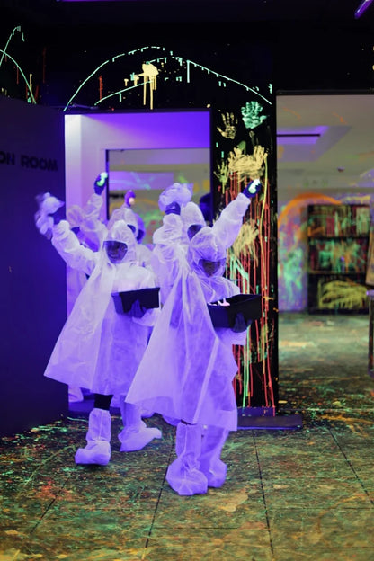 Children in protective suits hold glowing objects in a blacklight room with neon paint splatters.