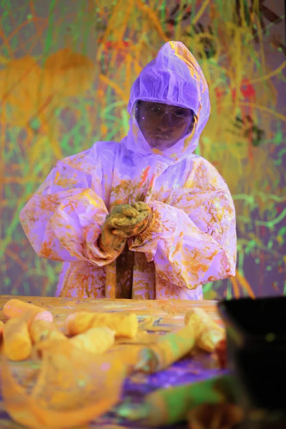 A person in a protective suit covered in paint stands in a colorful, abstract art studio.