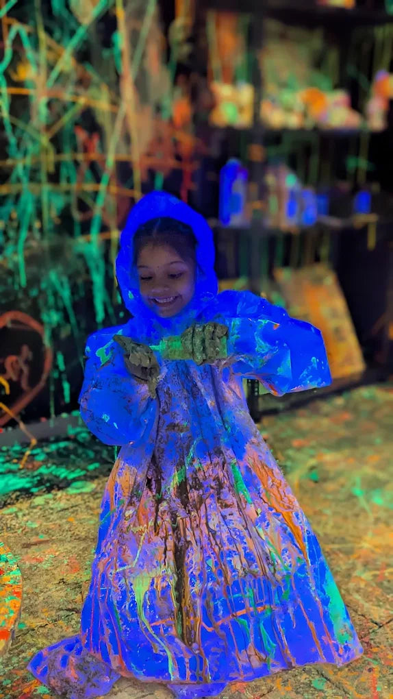 A young child in a blue poncho covered in neon paint smiles while holding a paint-covered object. The background is a dark, paint-splattered environment.