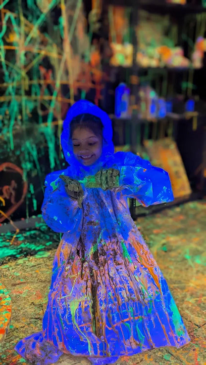 A young child in a blue poncho covered in neon paint smiles while holding a paint-covered object. The background is a dark, paint-splattered environment.