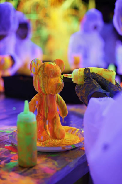 A person in gloves decorates a bear figurine with neon paint under blacklights.