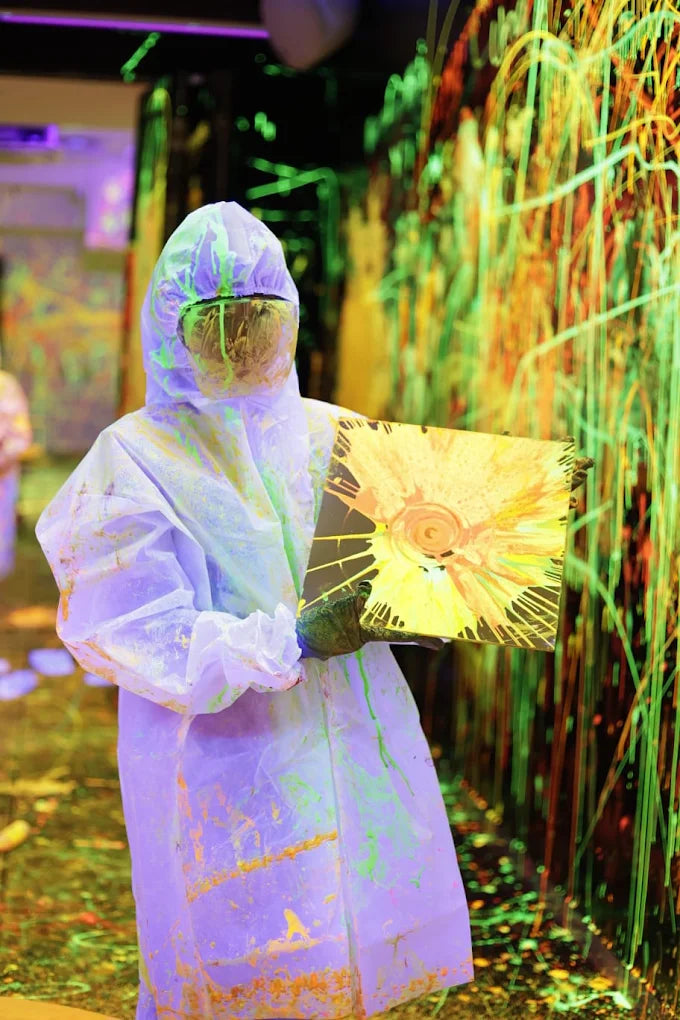 A person in a protective suit holds a splatter-painted canvas in a neon-lit room.