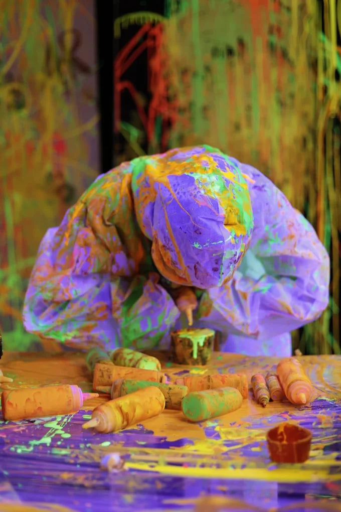 A person covered in purple and green paint is hunched over a table splattered with paint, surrounded by paint bottles.