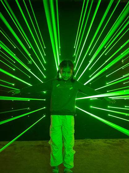 A young girl stands with arms outstretched amidst a dazzling display of green laser beams. The lasers radiate outwards from behind her against a dark background.