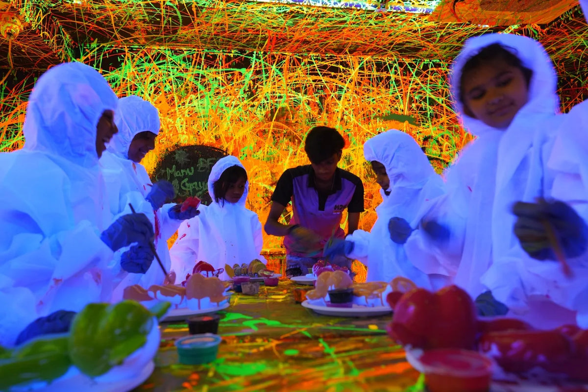 Children in protective suits paint at a table in a neon-lit room with a splattered background.