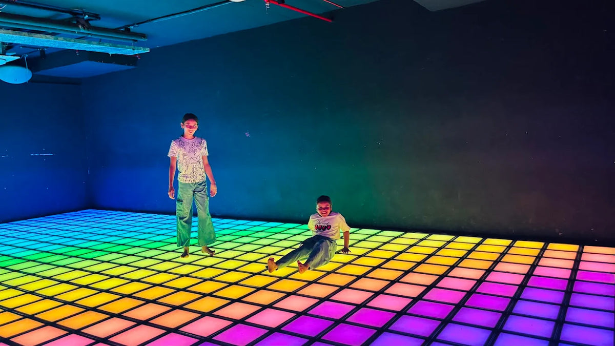 Two children play on a rainbow-colored light-up dance floor in a dark room.