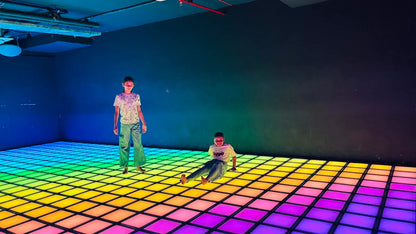 Two children play on a rainbow-colored light-up dance floor in a dark room.