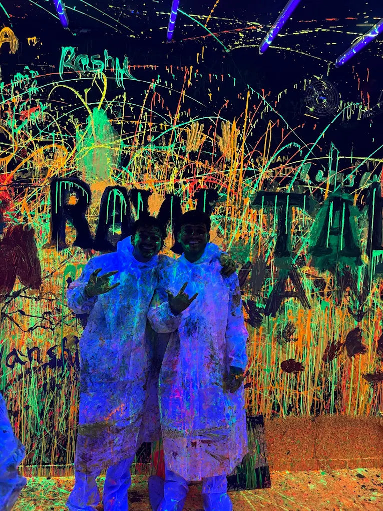 Two young adults in protective suits stand in a room splattered with neon paint under blacklights.