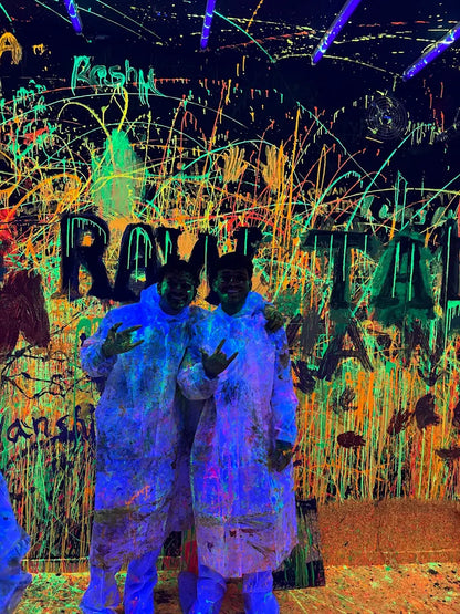 Two young adults in protective suits stand in a room splattered with neon paint under blacklights.