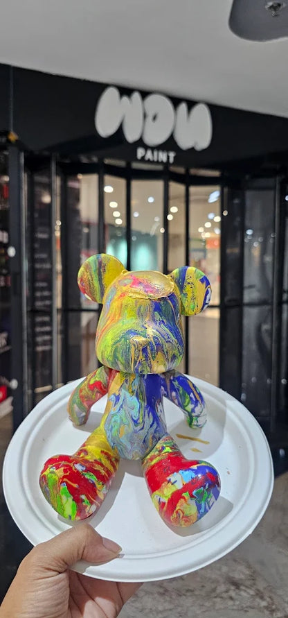 A colorful, paint-splattered bear figurine is held on a white plate in front of a "WOW PAINT" sign.