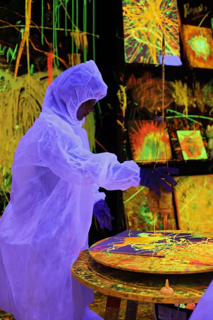 A person in a protective suit creates art with dripping paint under blacklights, surrounded by vibrant, glowing canvases.