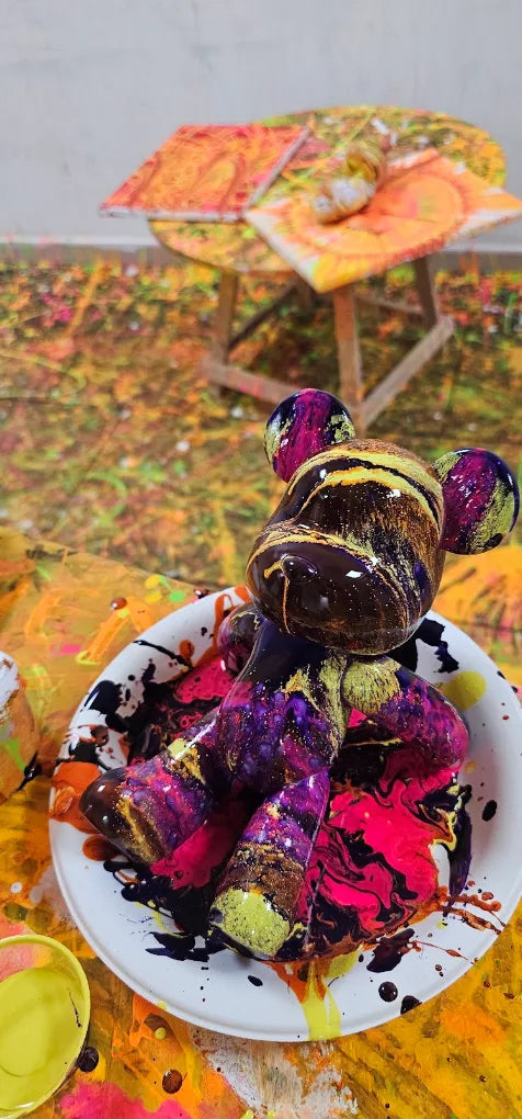 A colorful, paint-splattered teddy bear sits in a white bowl, surrounded by vibrant paint. The background is a chaotic canvas of bright, abstract art.