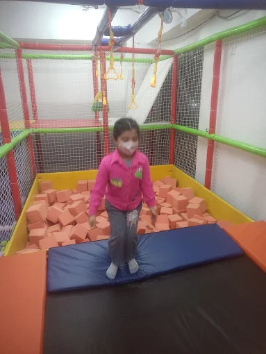 A young girl jumps on a trampoline in a foam pit. She is wearing a pink jacket and grey pants.