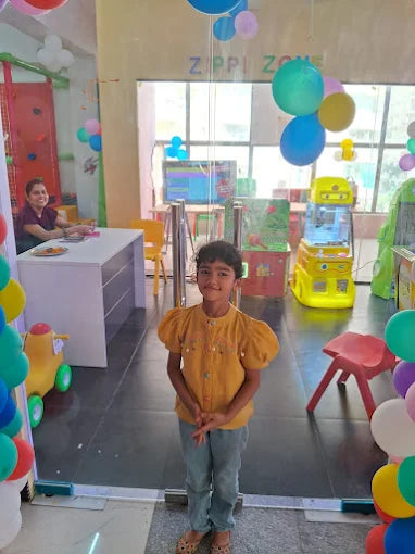 A young girl stands in a brightly decorated indoor play area with balloons and play equipment.