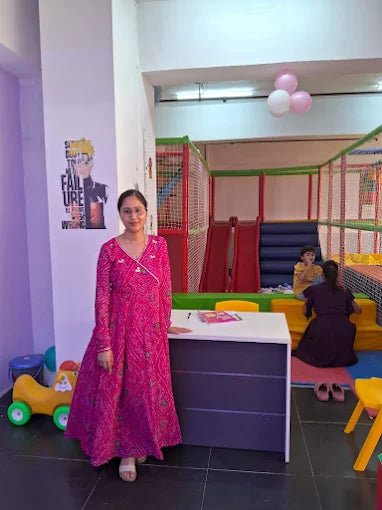 A woman in a pink dress stands in a play area with children in the background.