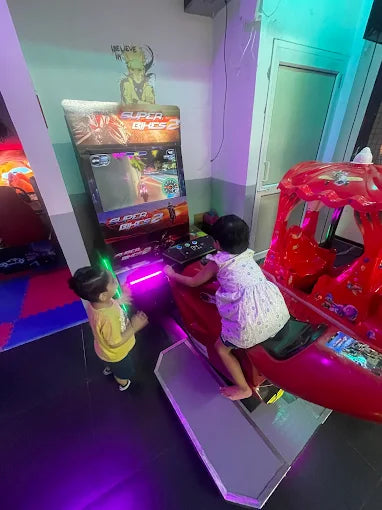 Two young children play a racing arcade game in a brightly lit entertainment center.