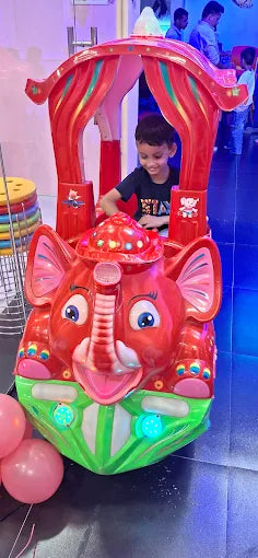 A young boy rides a red elephant-themed amusement park ride. Pink balloons are visible in the foreground.