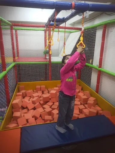 A young child in a pink shirt and jeans hangs from a monkey bar over a ball pit.