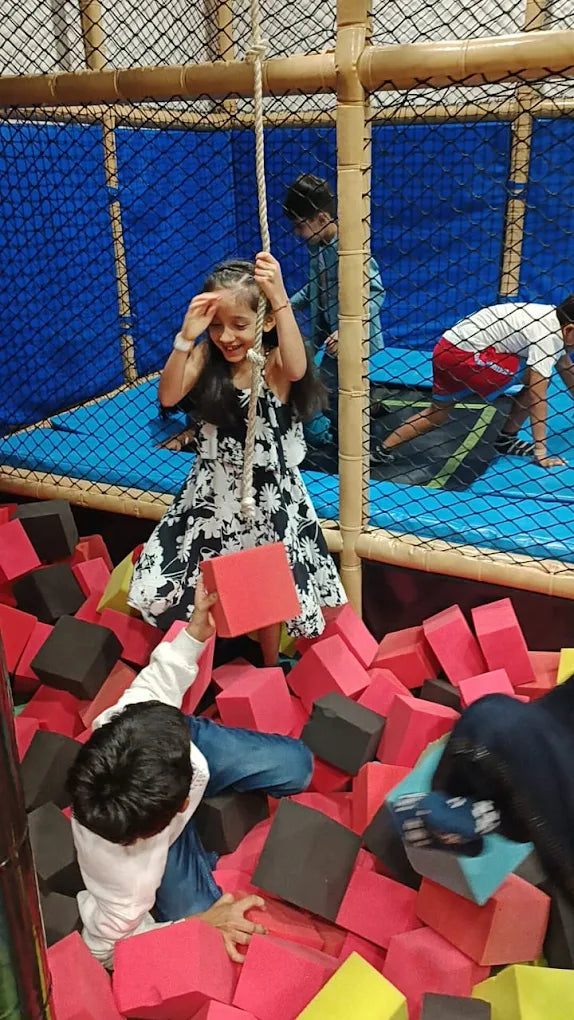 Children play in a foam pit and on a trampoline-like structure. A girl swings from a rope while a boy reaches for a foam block.