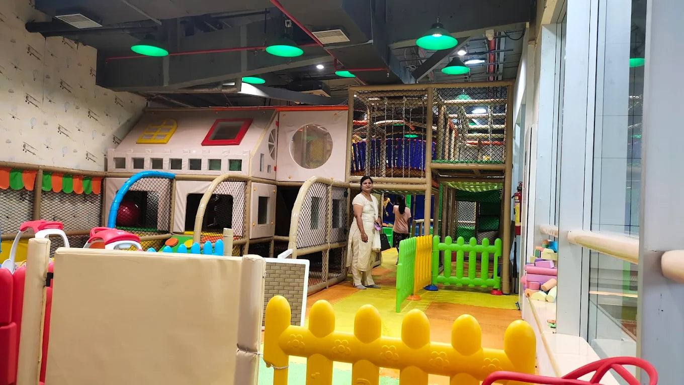 A woman stands in a brightly lit indoor playground with colorful structures and soft flooring.