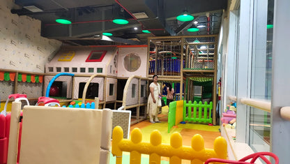 A woman stands in a brightly lit indoor playground with colorful structures and soft flooring.