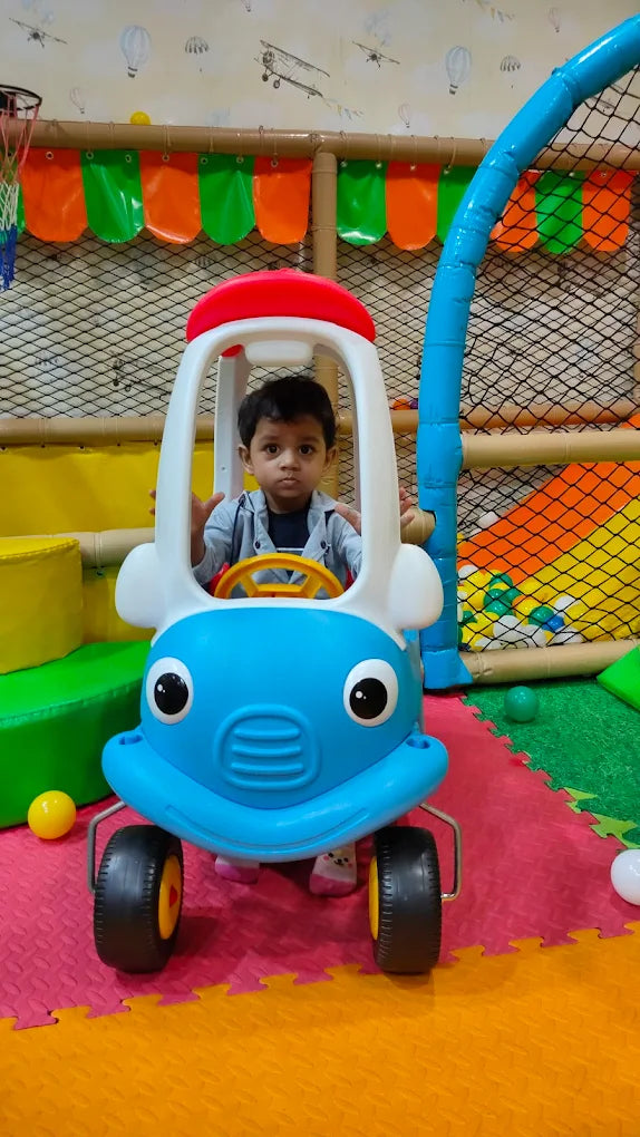 A young child sits in a blue toy car in a colorful indoor playground. The child is looking forward with hands on the steering wheel.