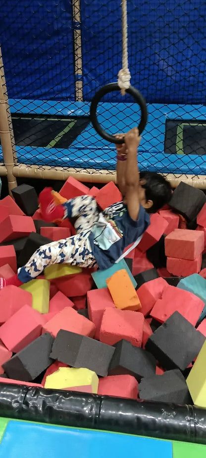 A child hangs from a ring over a foam pit filled with colorful blocks.