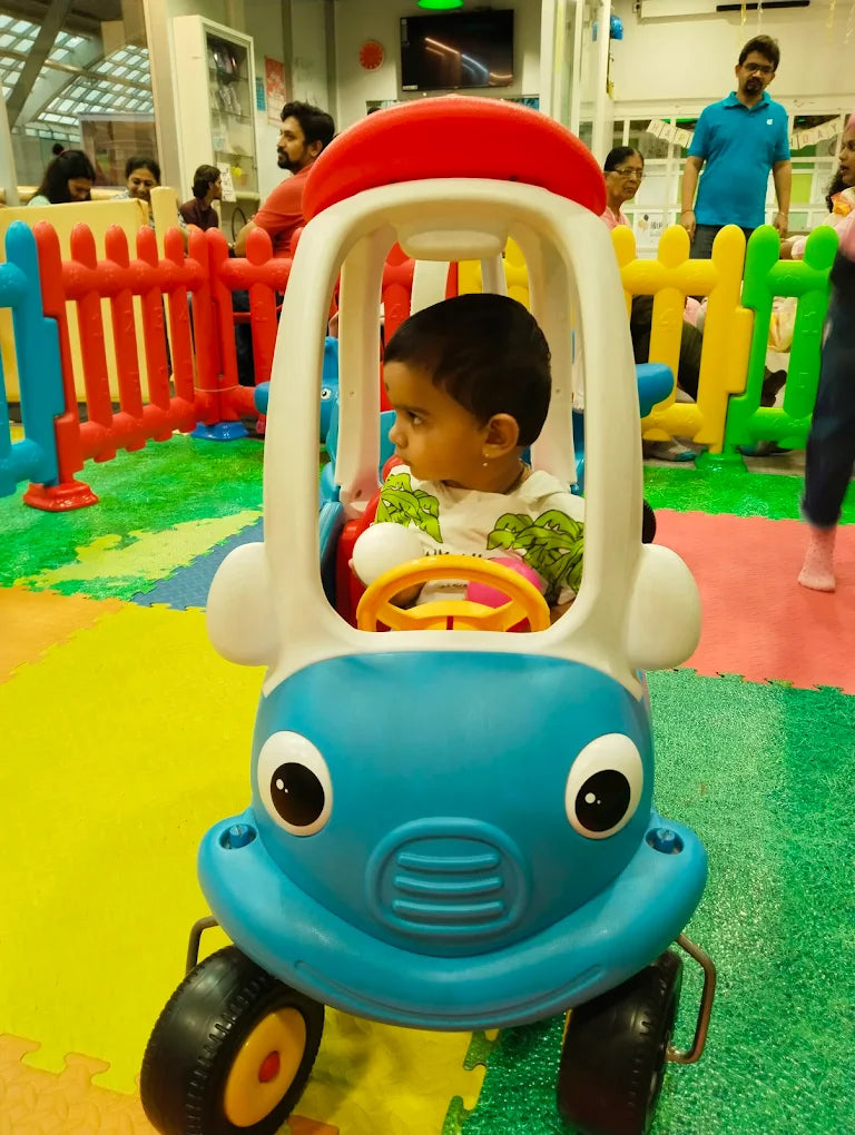 A toddler sits in a toy car at an indoor playground. Other children and adults are visible in the background.