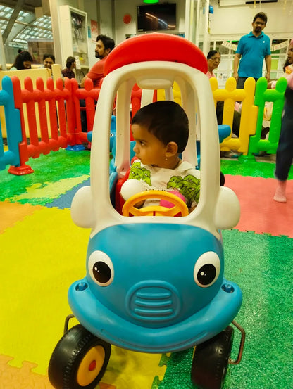 A toddler sits in a toy car at an indoor playground. Other children and adults are visible in the background.