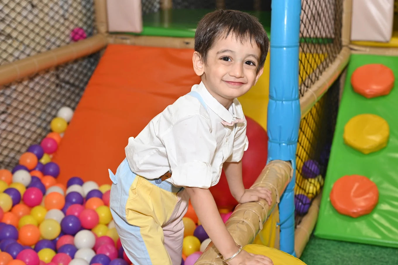 A young boy smiles while playing in a colorful ball pit. He is wearing a white shirt and pastel overalls.
