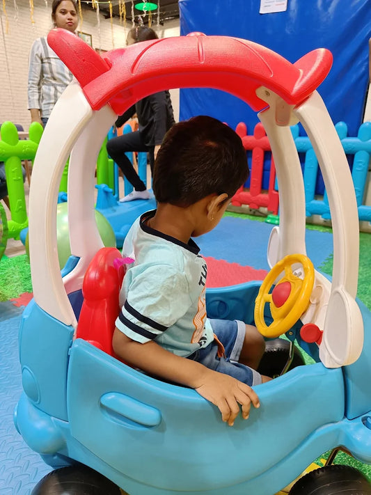 A young child sits in a toy car at an indoor playground.