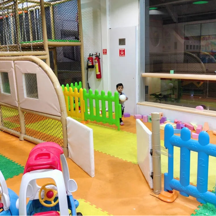 A young child holds a ball in a colorful indoor playground with soft flooring and play structures.