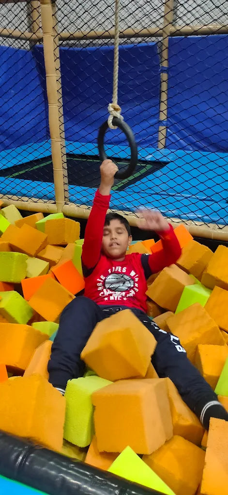 A young boy in a red shirt hangs from a ring in a foam pit at a trampoline park.