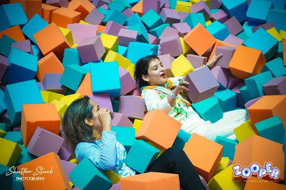 Two people playing in a pit filled with colorful foam cubes, with 'Zoopa' branding visible.