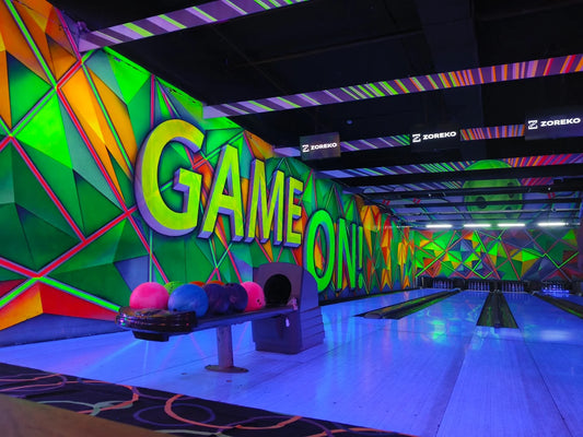 A neon-lit bowling alley with colorful geometric murals and bowling balls on a rack.