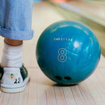 Person wearing blue jeans and white sneakers standing next to a blue bowling ball on a bowling lane.